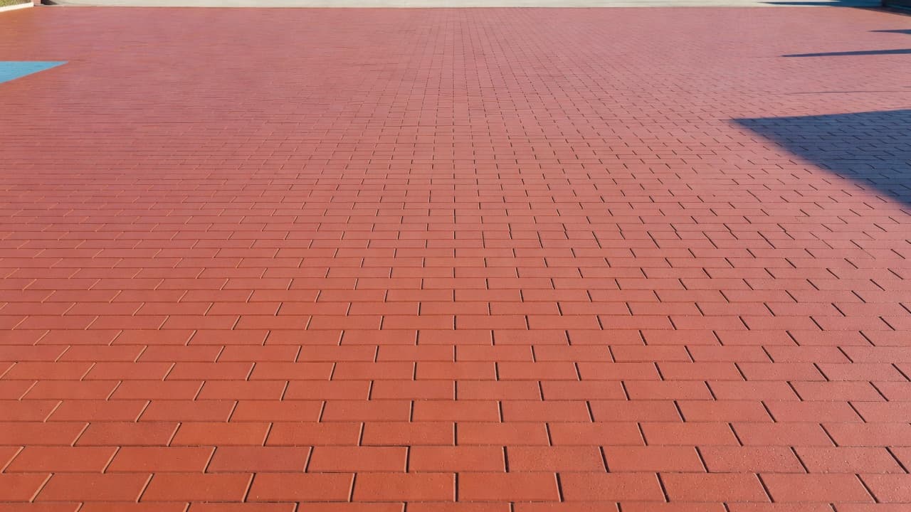 A pristine red brick driveway stretches toward a modern home, its surface sealed to a smooth, clean finish. Subtle blue accents line the edges of the vibrant, sun-drenched masonry path.